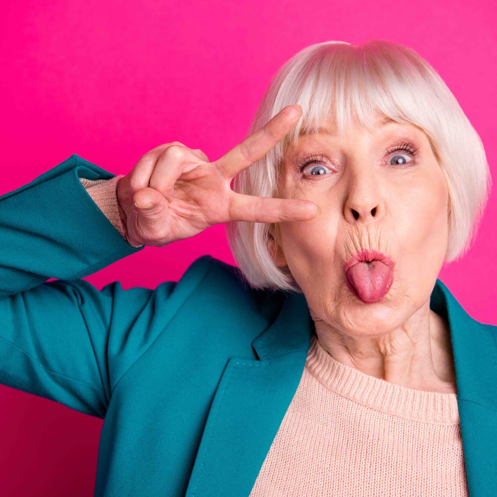 Close-up portrait of nice attractive cheerful funky gray-haired lady wearing blue jacket showing v-sign near eye tongue out fooling isolated on bright vivid shine vibrant pink fuchsia color background